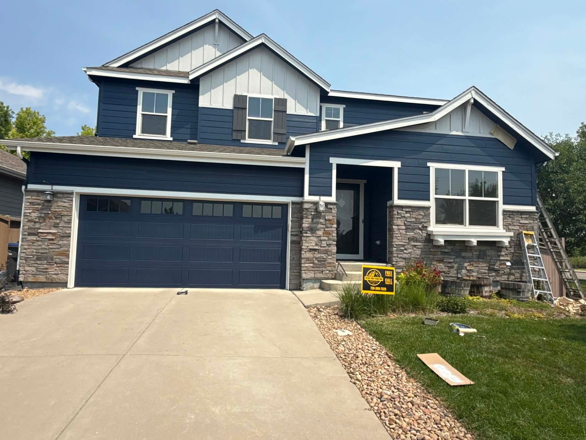 A two-story blue house with stone veneer, a garage, and a light-colored front entrance under a clear blue sky.
