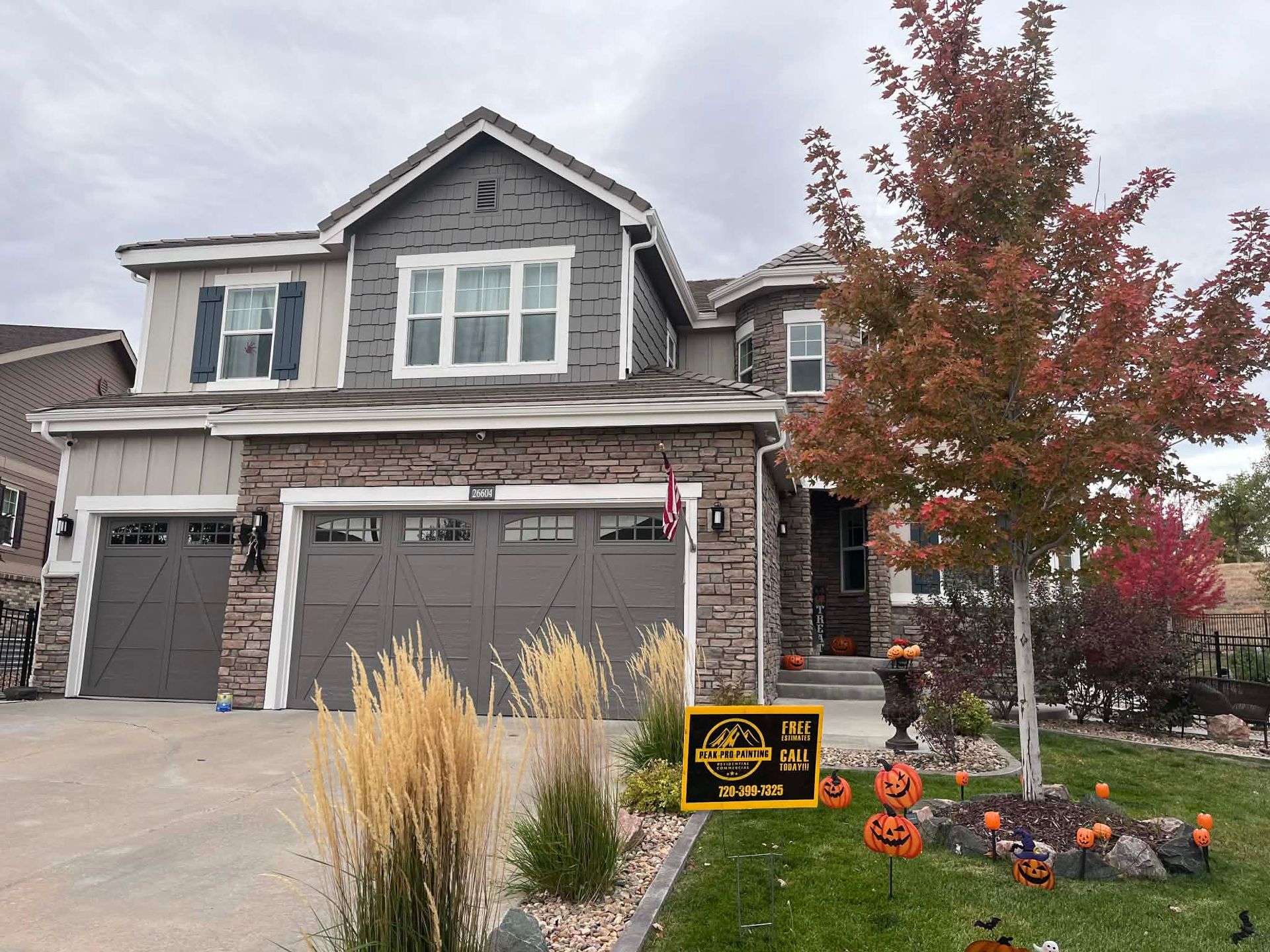 A two-story suburban home with stone siding and dark garage doors decorated with Halloween pumpkins and a yard sign.