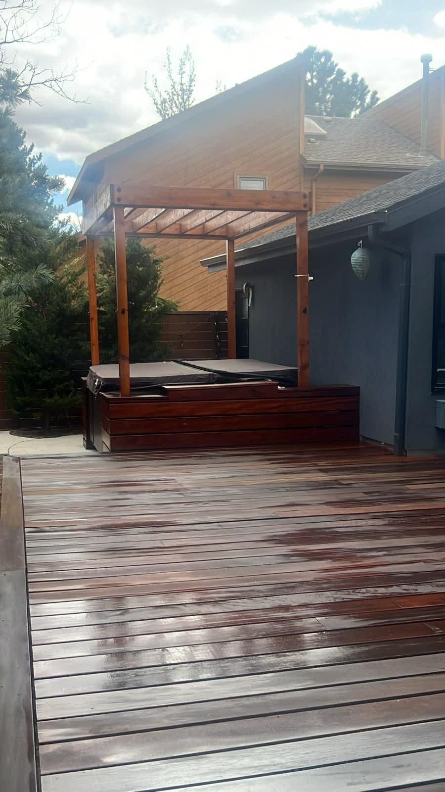 A wet wooden deck leads to a hot tub enclosed by a dark wood platform and a pergola, set against a house and pine trees.