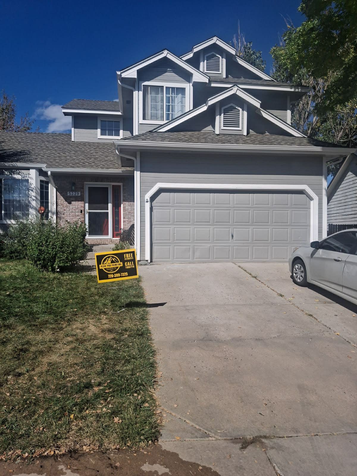 A two-story grey suburban house with a two-car garage, a concrete driveway, and a small yard under a blue sky.