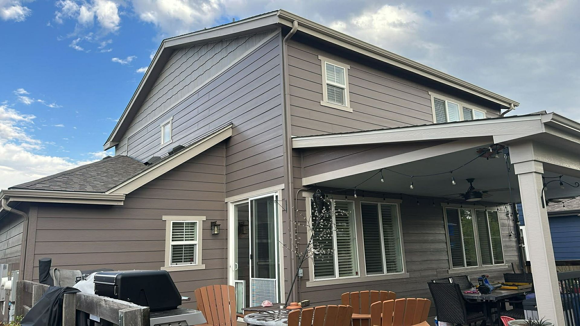 A two-story brown house with a covered patio, outdoor furniture, and a grassy backyard under a cloudy sky.