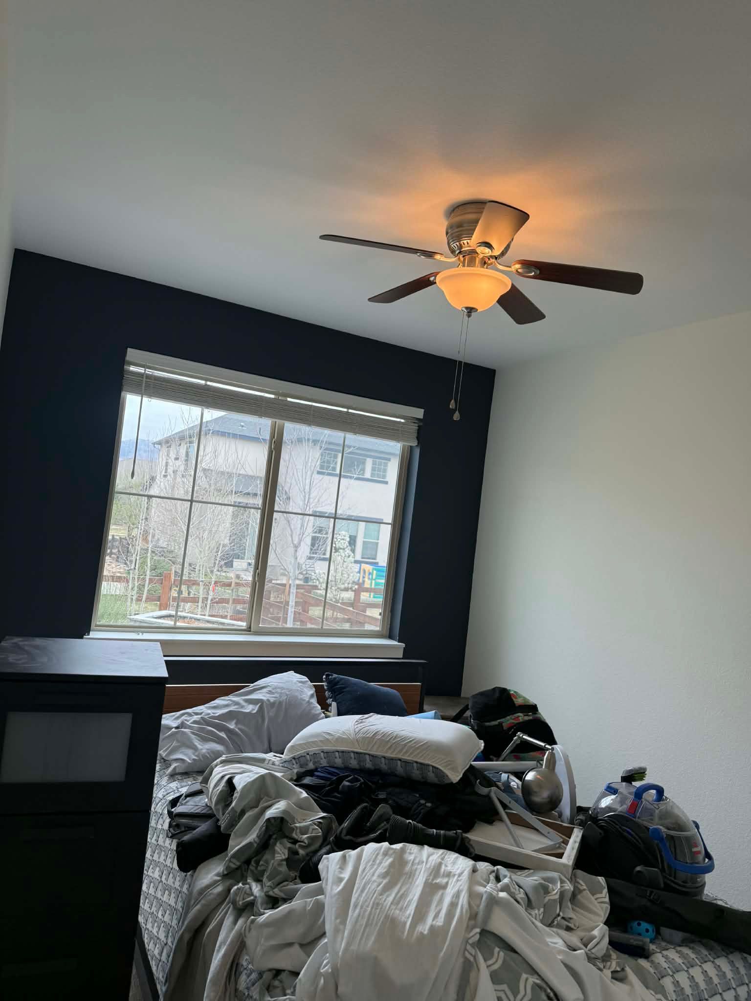 A bedroom with a dark blue accent wall, a window, a ceiling fan, and a messy bed with pillows and scattered items.