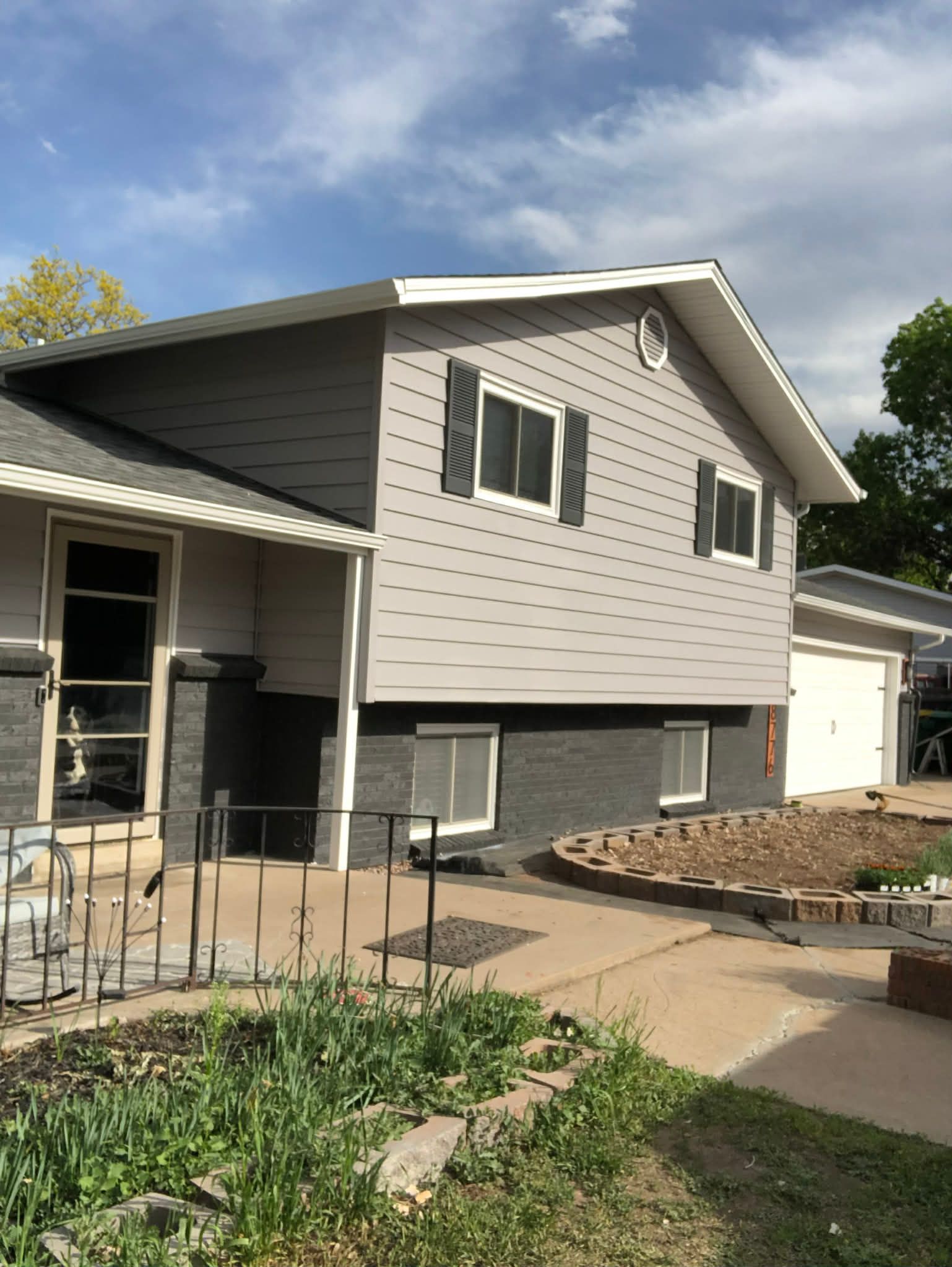 A split-level house with light gray siding, a dark gray brick foundation, two windows with shutters, and a concrete patio.