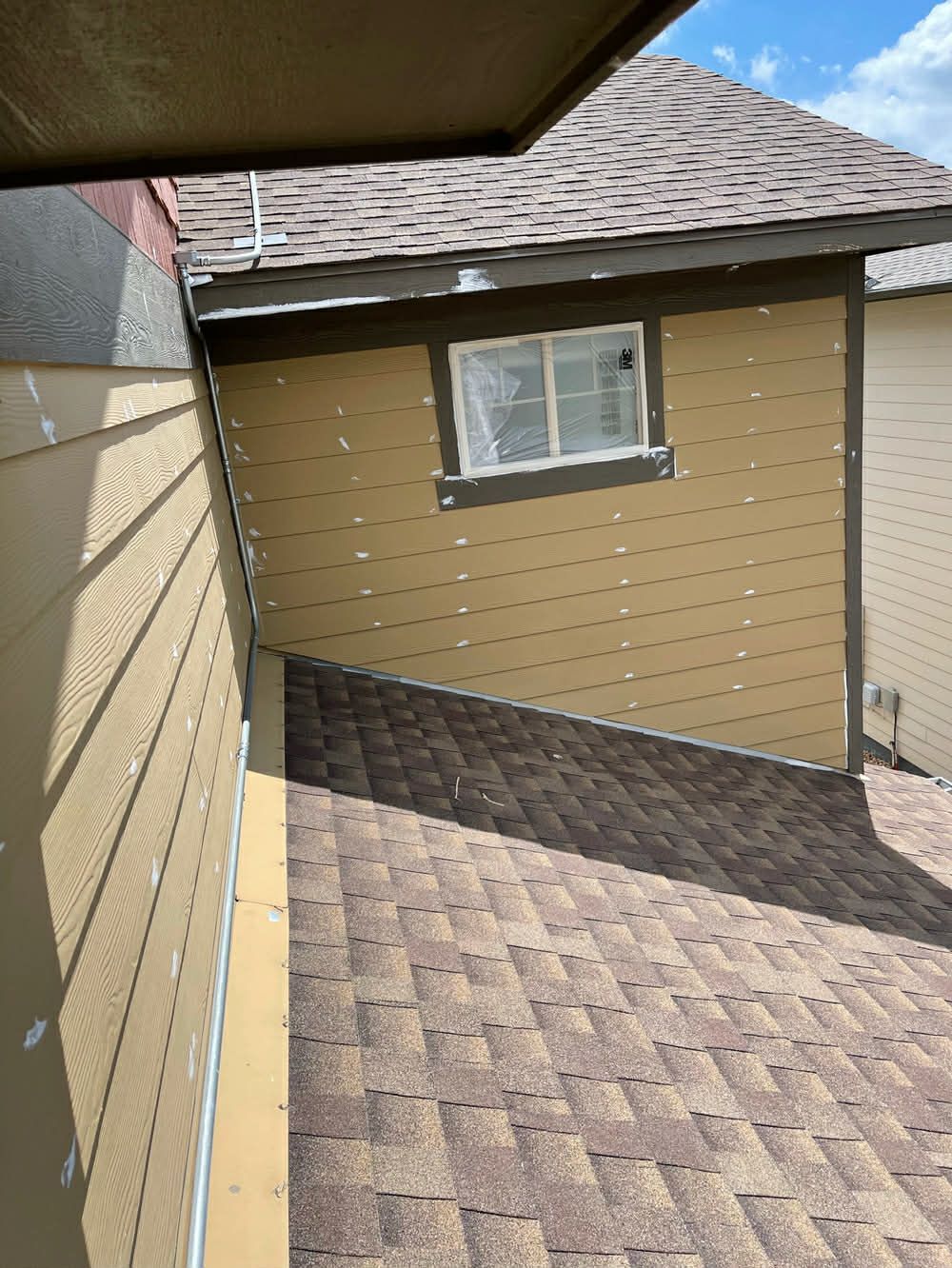 An angled view of a house exterior showing tan horizontal siding and a dark brown shingled roof sloping toward the wall.