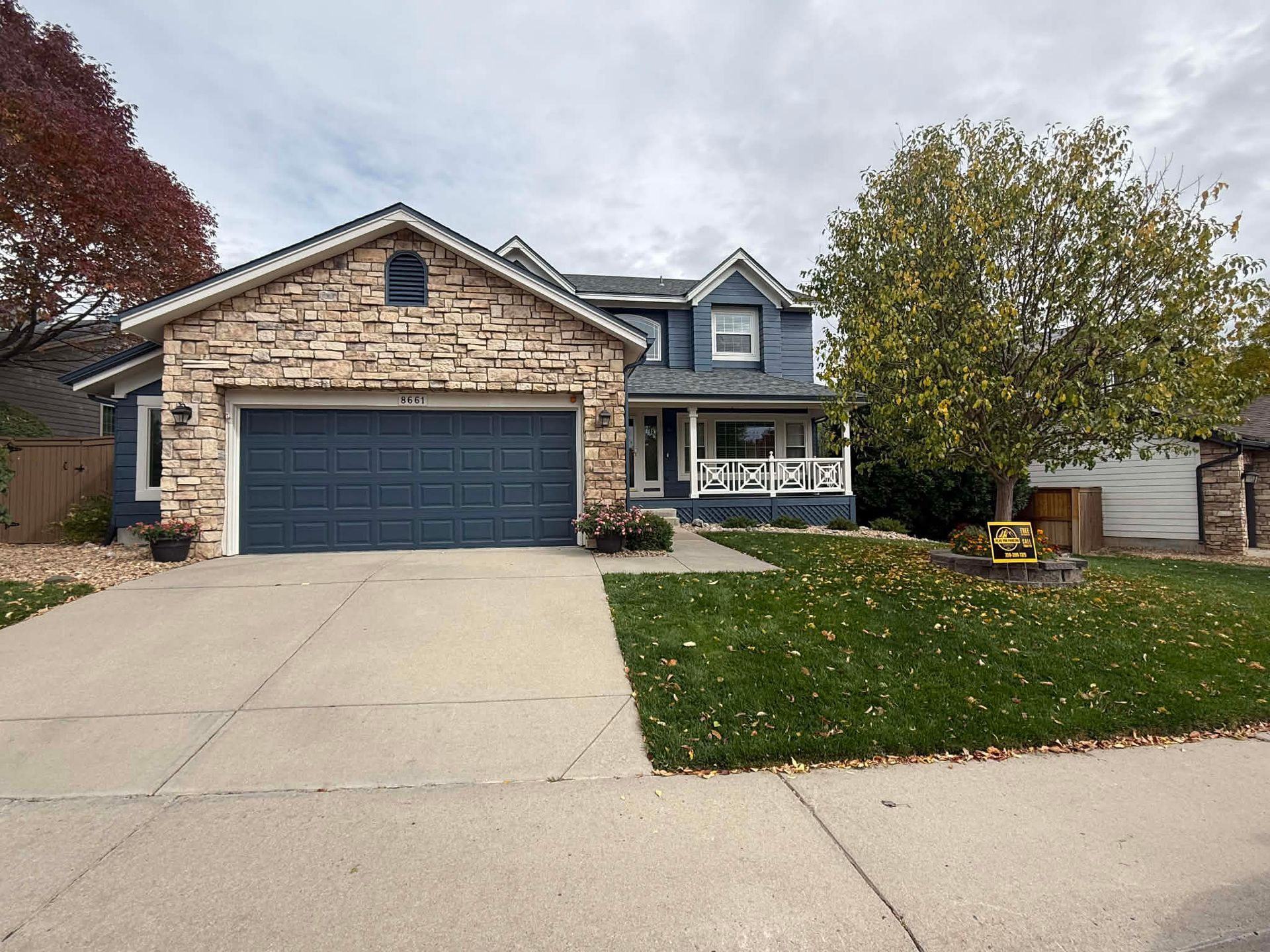 A two-story blue house with a stone facade, a two-car garage, and a front lawn with a tree under an overcast sky.