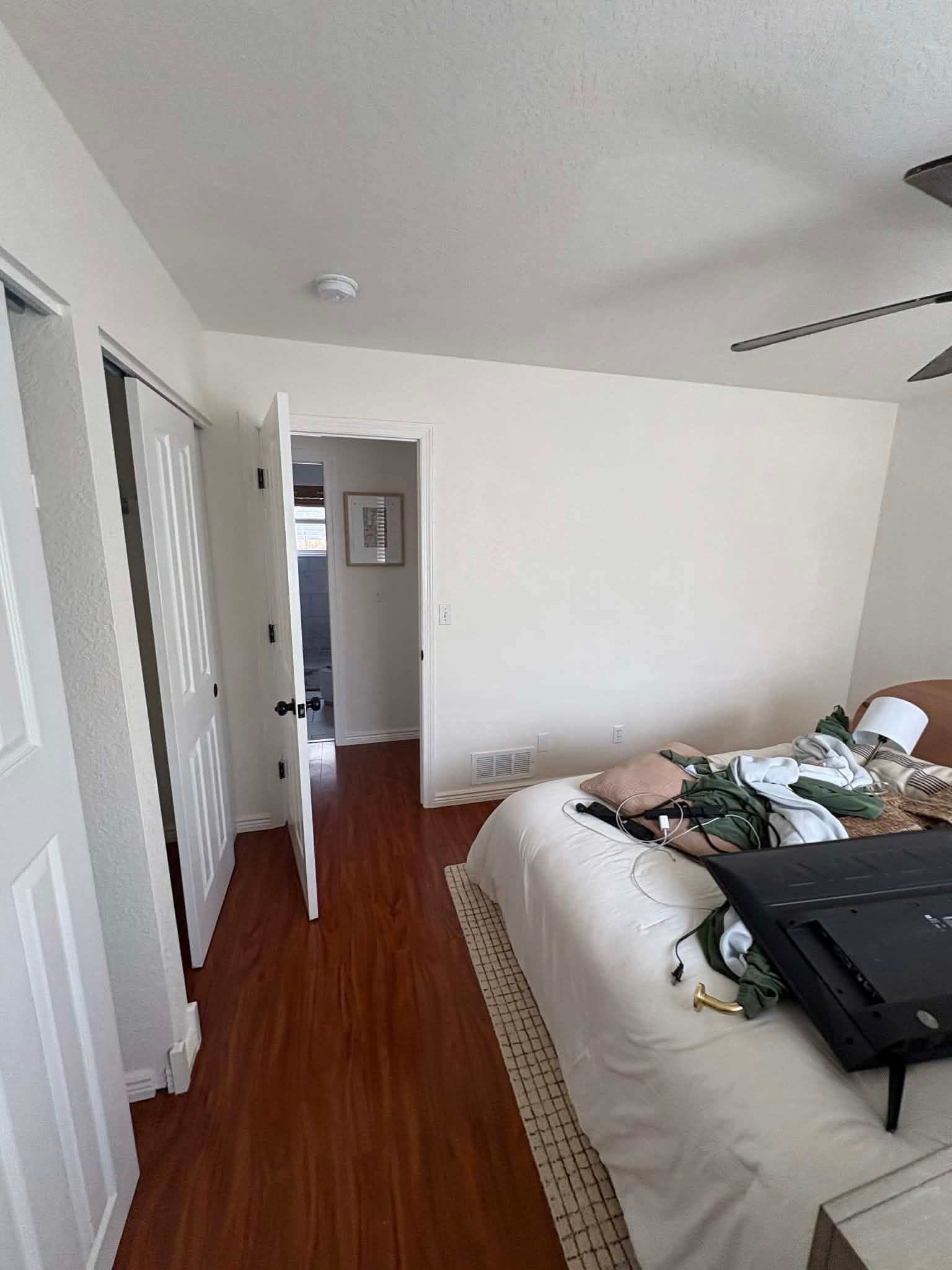 A bedroom featuring wood-look floors, a white bed with a black TV on top, and an open doorway leading to a hallway.