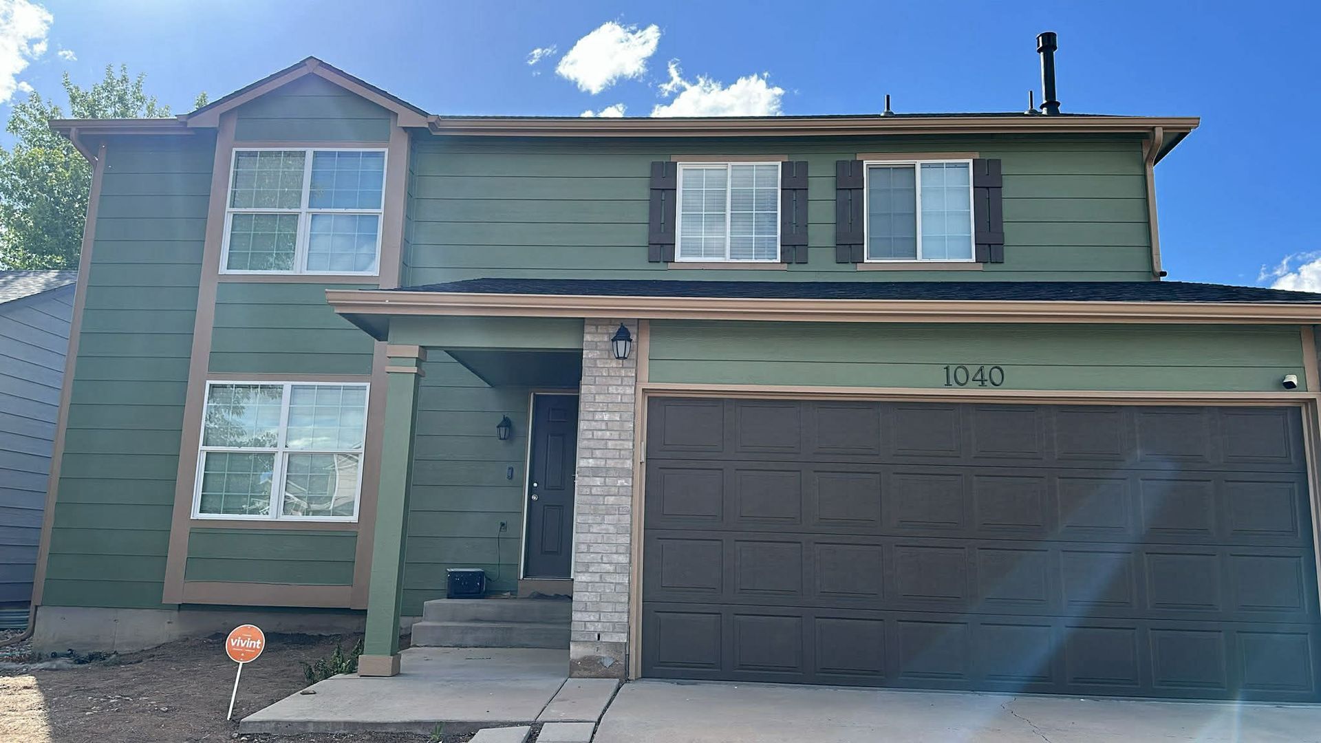 A two-story house with olive green siding, a brown garage door, stone accents, and several rectangular windows.