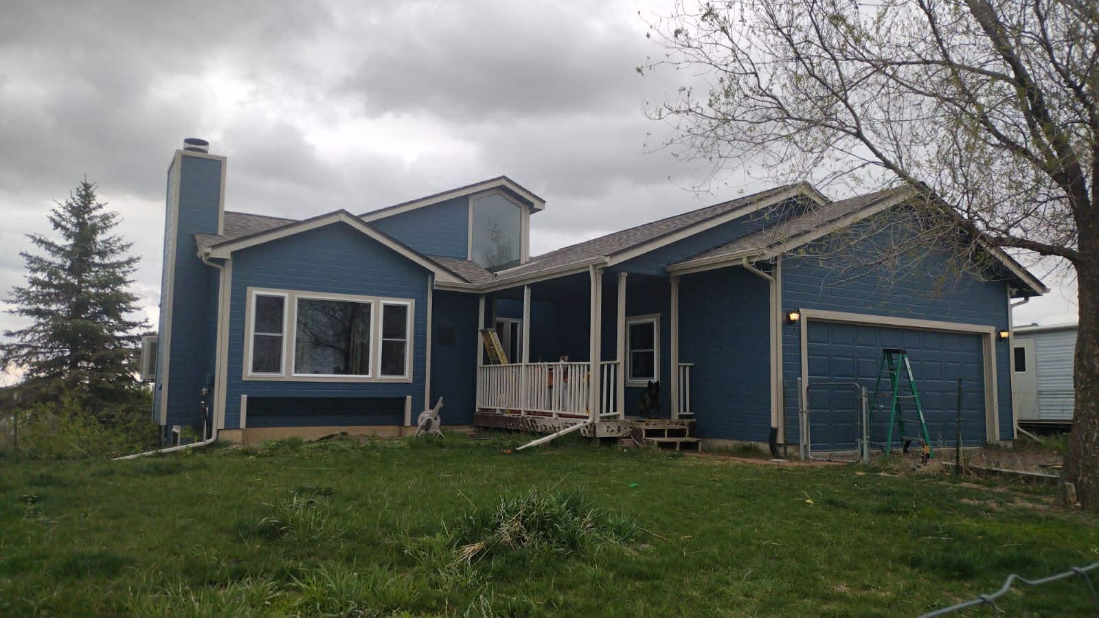 A blue house with a prominent chimney, front porch, and garage, set against a cloudy sky with green grass in the yard.