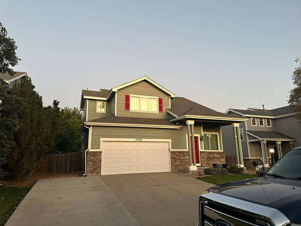 A two-story suburban house with olive green siding, a two-car garage, and a porch, viewed from a residential driveway.