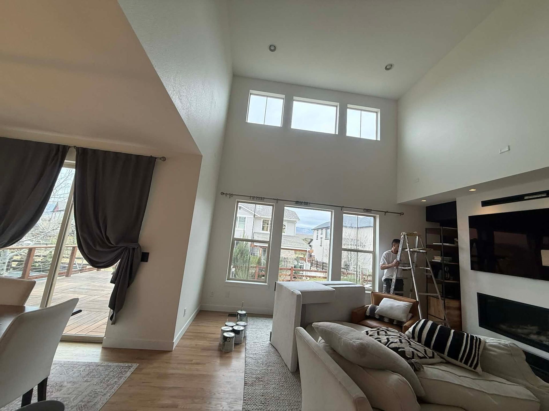 A high-ceilinged, modern living room with large windows, neutral furniture, and a person working on a ladder.