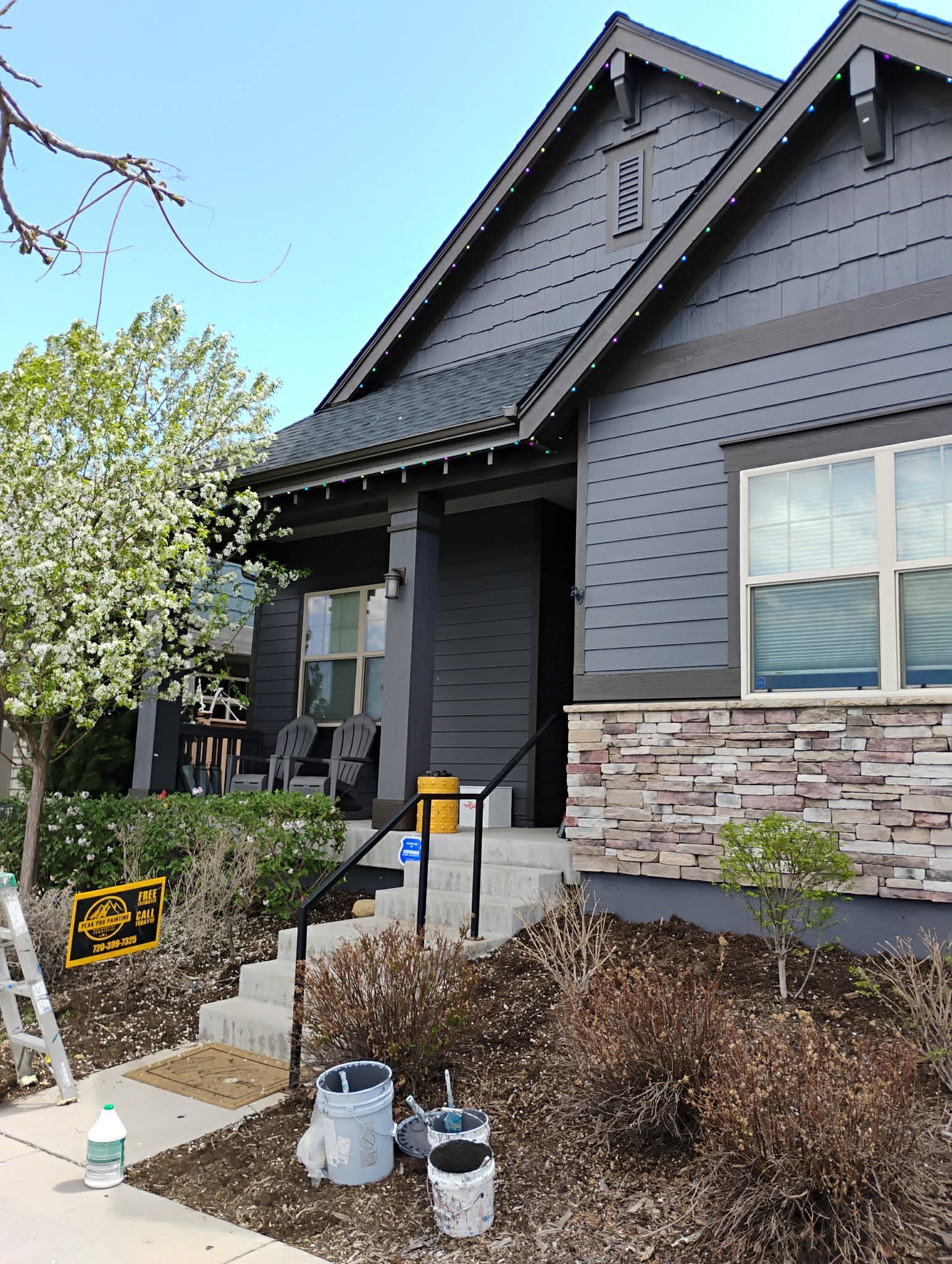 A grey, single-story house with stone veneer siding, a front porch, and construction supplies sitting in the front yard.