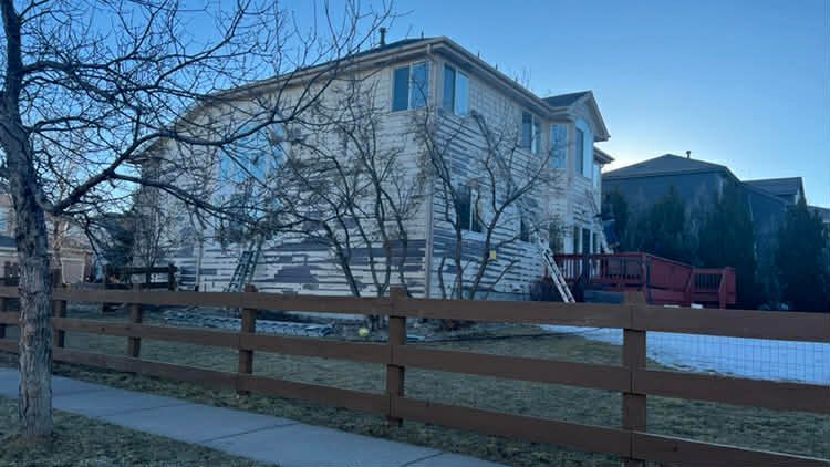 A two-story beige house sits behind a wooden post-and-rail fence on a sunny day with bare trees in the foreground.