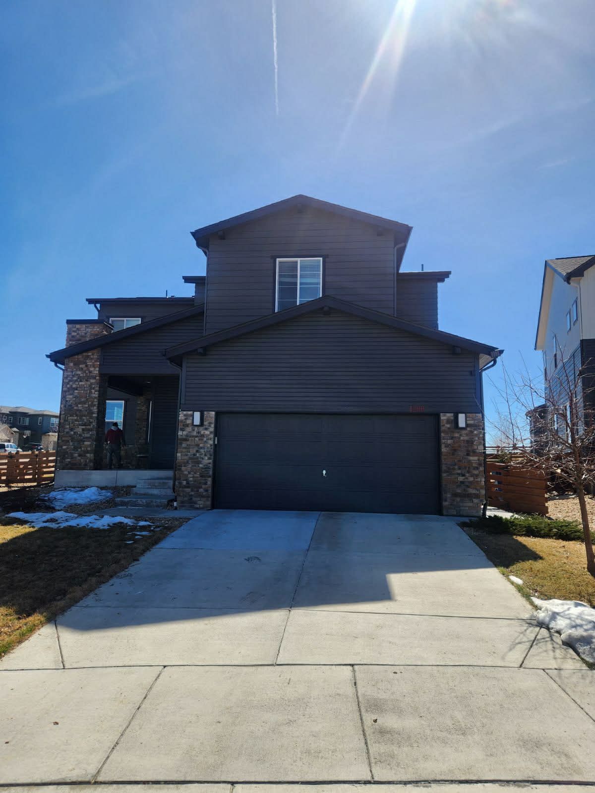 A two-story dark brown house with a large garage, stone siding accents, and a concrete driveway under a clear blue sky.