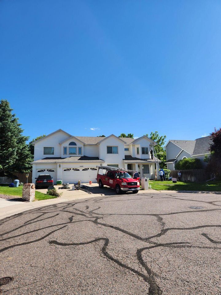 A suburban house with a white exterior, a red service truck in the driveway, and a cracked asphalt street in front.