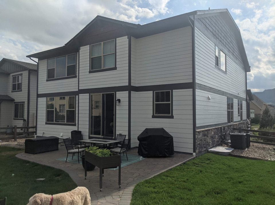 A white two-story house with dark trim and a stone base, featuring a backyard patio with furniture and a grill.