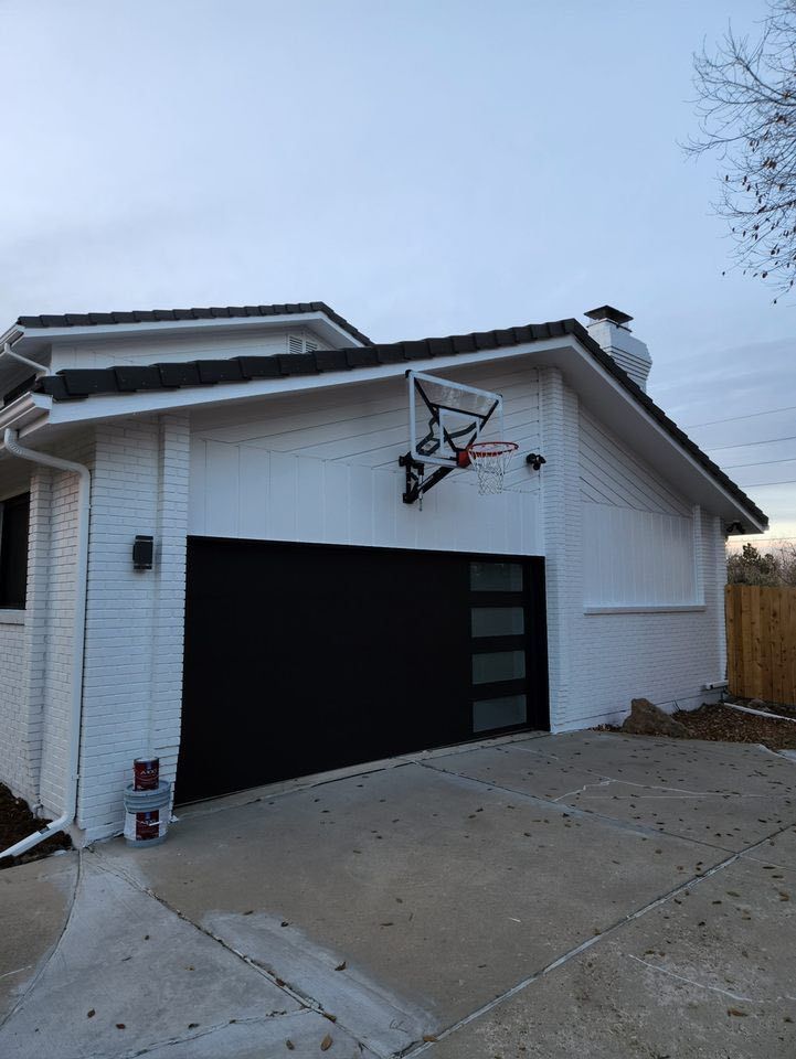 A white house with a dark garage door and a basketball hoop mounted above it, set against a twilight sky.