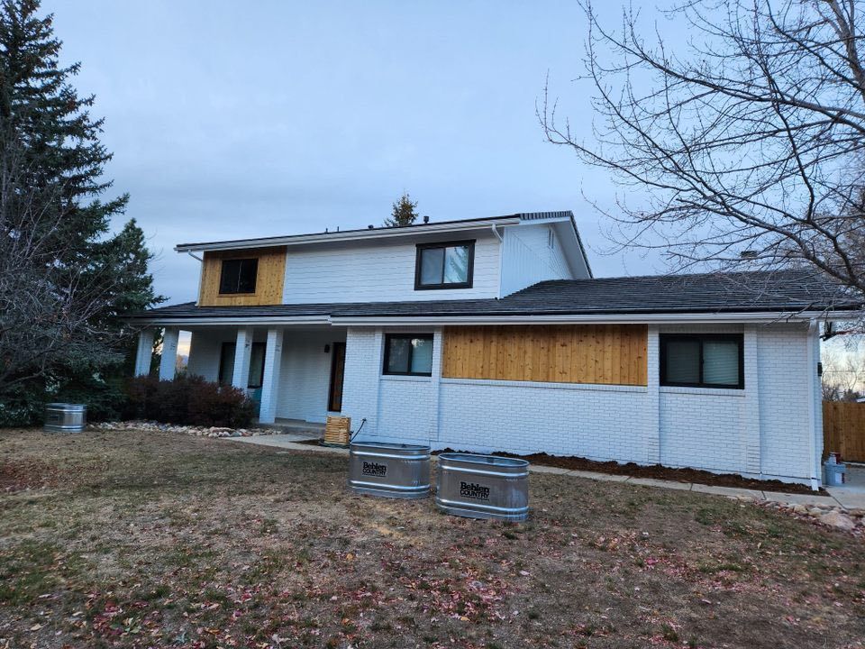 A two-story white house with wood siding patches under windows, featuring a front porch and two metal bins in the yard.