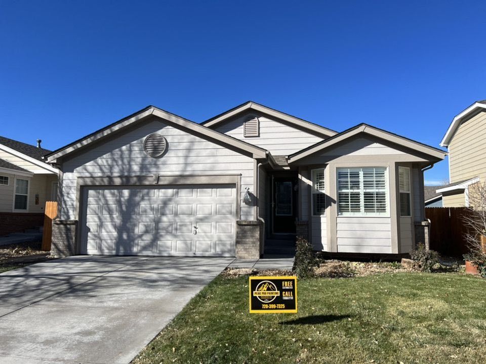A single-story suburban house with beige siding, a two-car garage, and a real estate sign on the front lawn.