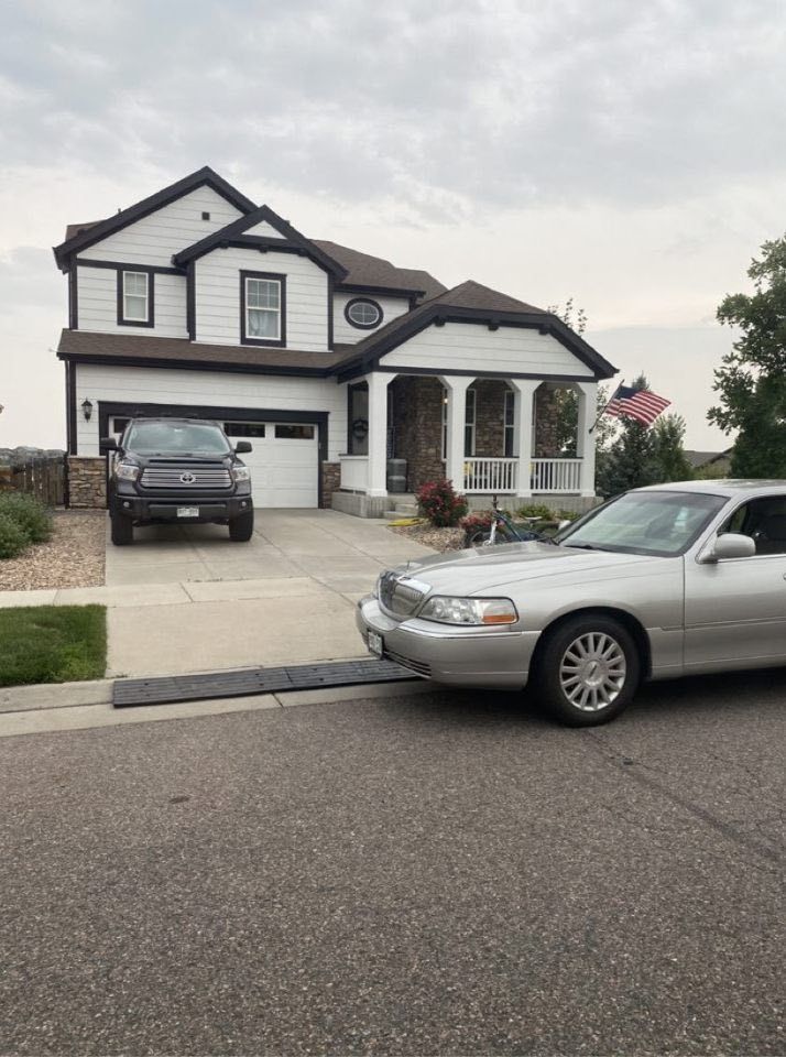A two-story house with a driveway, a parked truck, and a silver sedan driving over a rubber curb ramp.