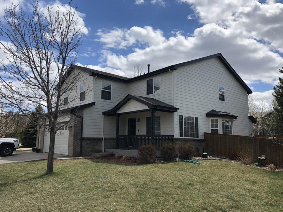 Two-story, light gray suburban house with a dark roof, a front porch, and an attached garage under a partly cloudy sky.