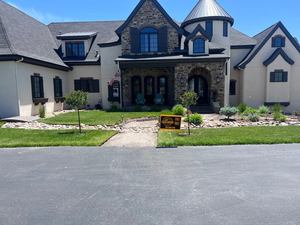 A large two-story house with stone and stucco exterior, a turret, and a lawn with a yellow real estate sign in the yard.