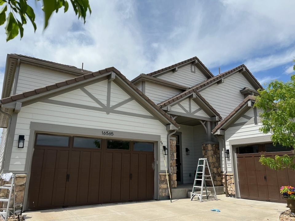 A two-story suburban home with light siding, dark wood garage doors, stone accents, and a ladder by the front entrance.