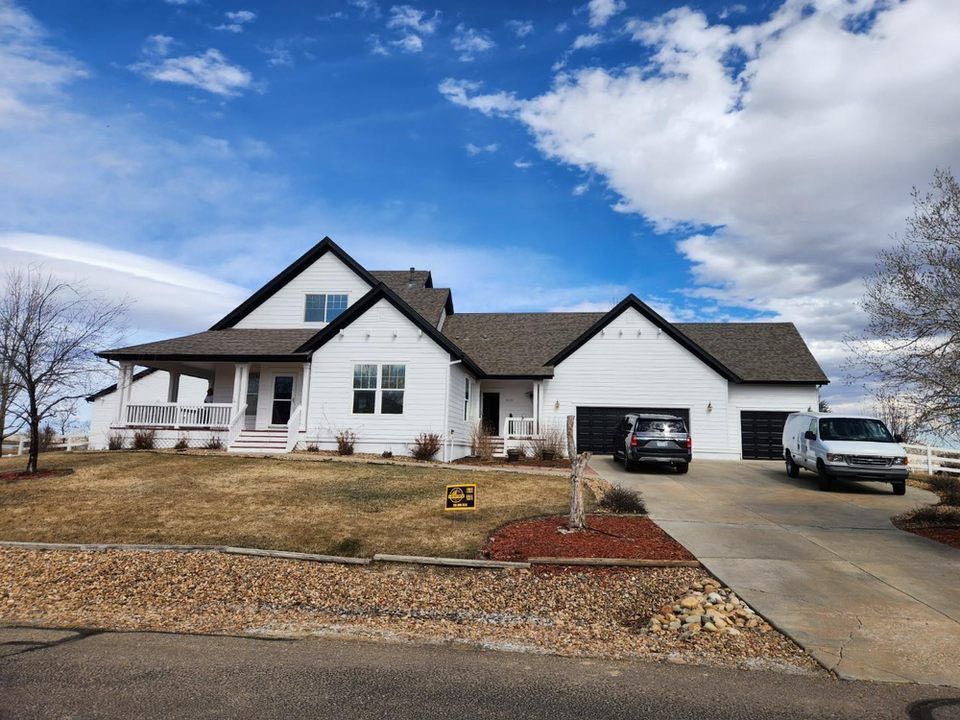 A white two-story farmhouse-style home with black trim, a wrap-around porch, and a large garage on a sunny, cloudy day.