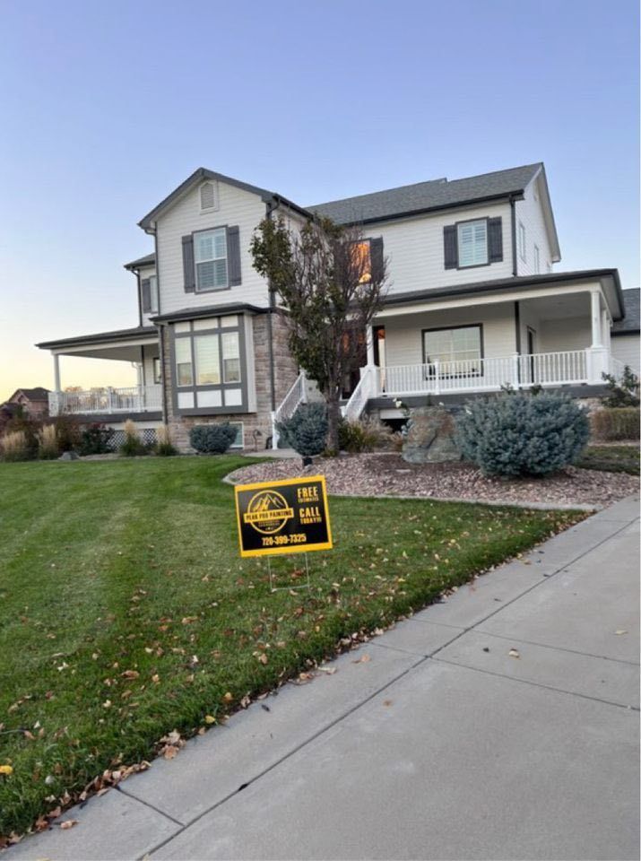 A two-story house with a porch and a white exterior sits behind a manicured lawn with a yard sign and blue shrubs.