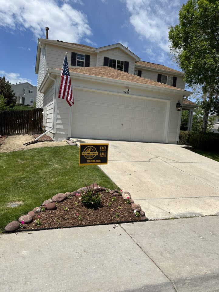 A two-story light-colored suburban house with a two-car garage, an American flag, and a yard sign on a sunny day.