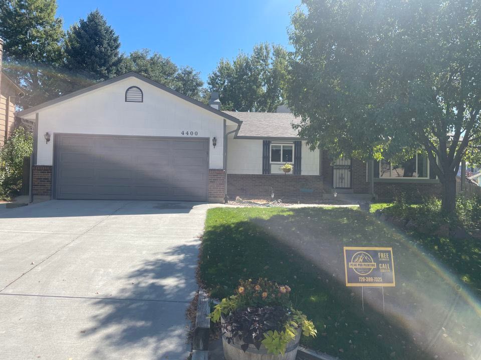 A single-story suburban house with a large garage, light-colored siding, a brick base, and a small sign on the front lawn.
