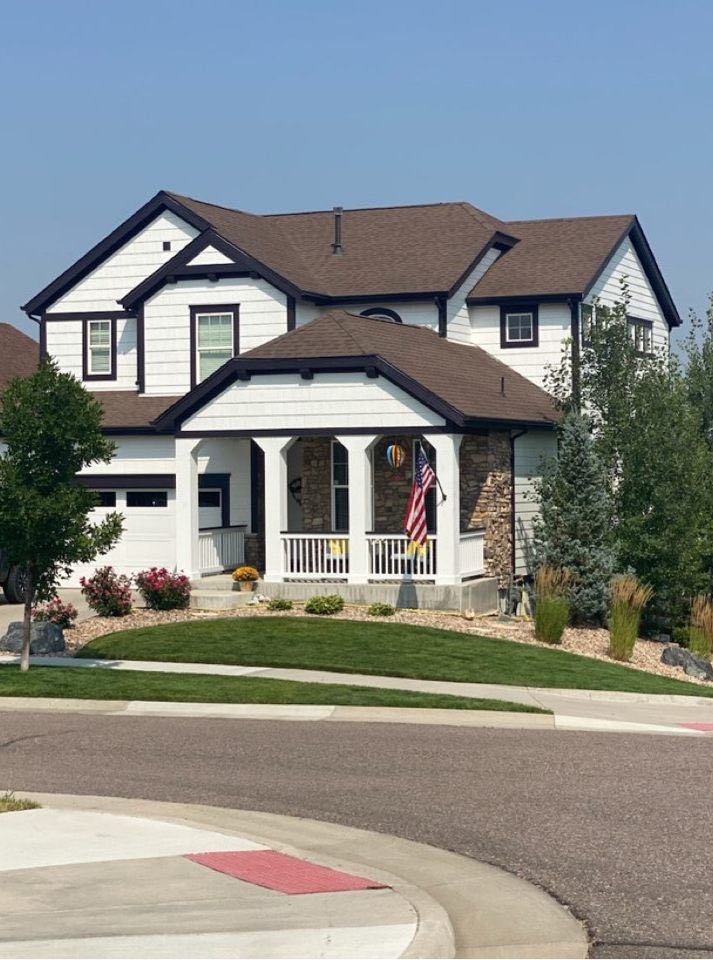 A two-story white house with dark trim, stone accents on the front porch, and a brown shingled roof under a clear sky.