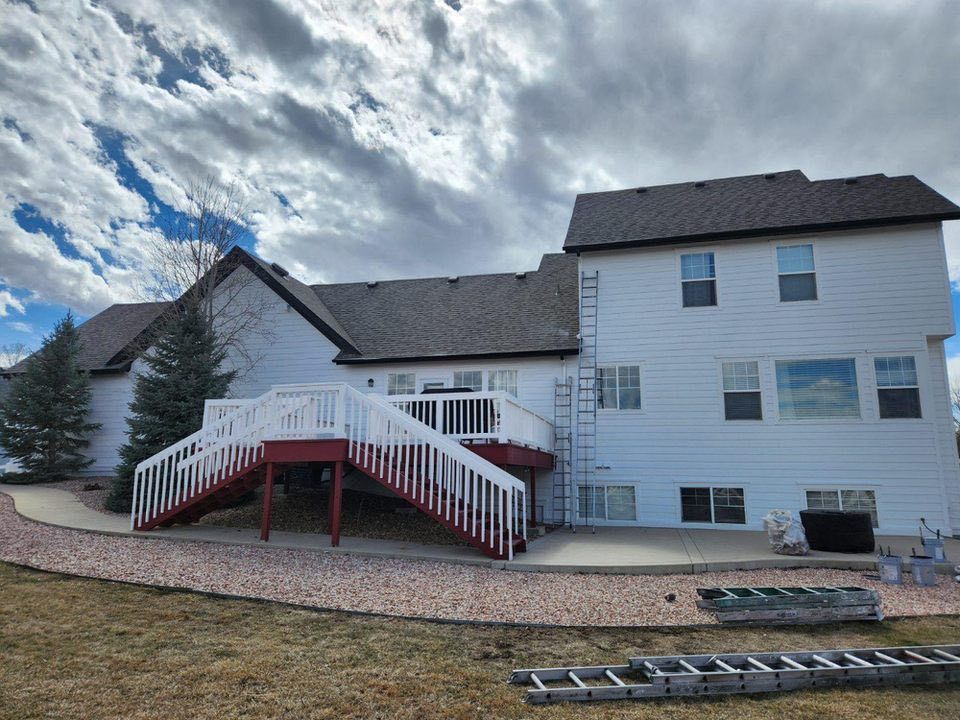 A two-story white house with a deck, red stairs, and a gravel patio under a cloudy sky.