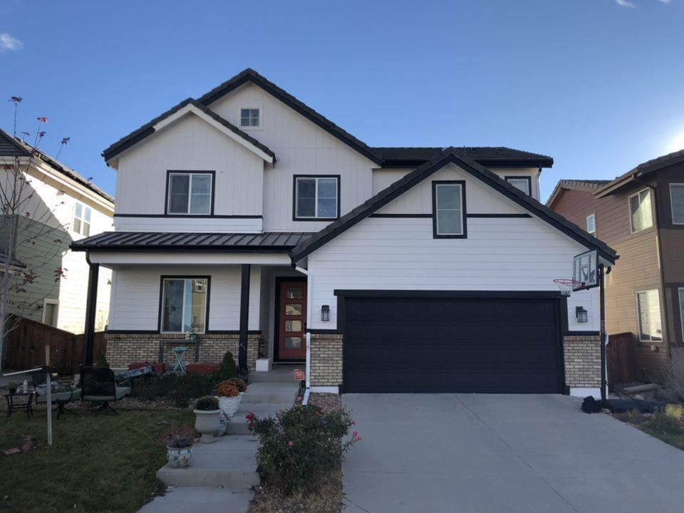 A two-story house with white siding, dark trim, a stone foundation, a black roof, and a two-car garage under a blue sky.