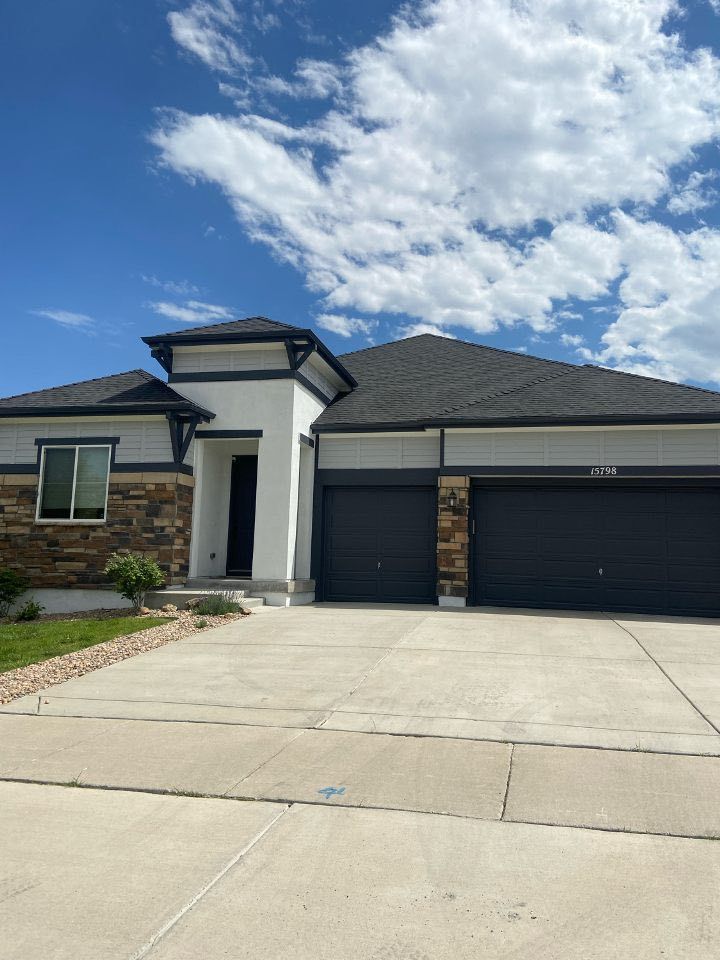 A single-story suburban house with white stucco, stone accents, a dark roof, and a two-car garage under a blue sky.