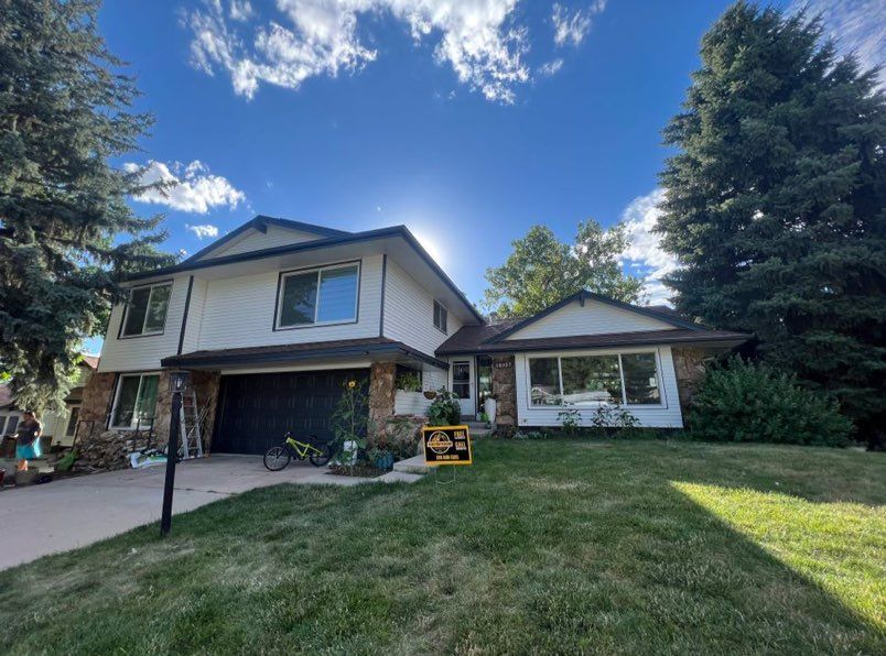 A two-story house with cream siding and dark trim, a black garage door, and a front lawn under a blue, sunny sky.