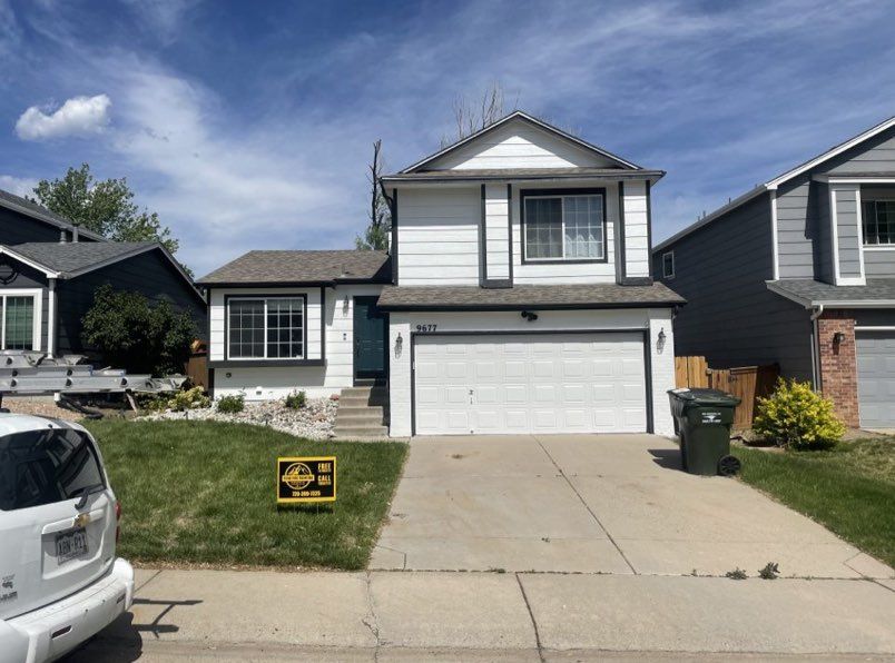 A two-story house with white siding and gray trim, featuring a garage, a small front lawn, and a green trash bin.