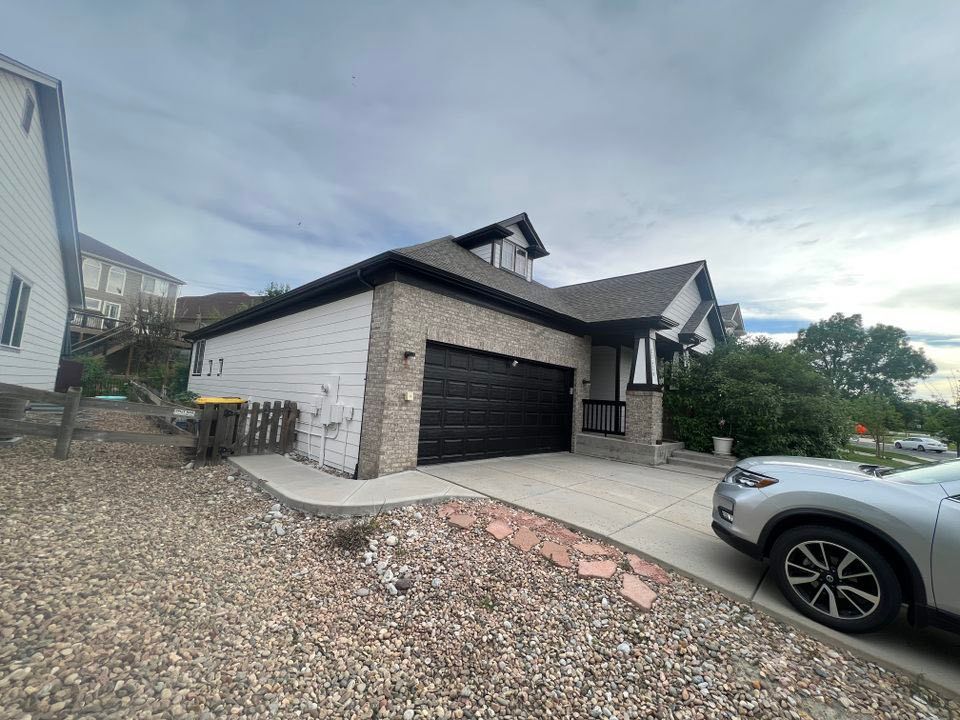 A house with a beige brick exterior, black garage door, and white siding on the side, next to a car in the driveway.