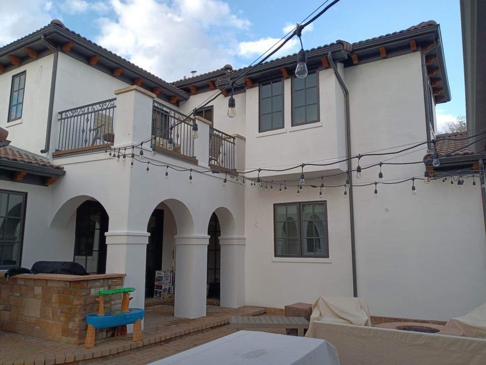 A two-story white stucco home exterior with a balcony, arched patio columns, and hanging string lights over a patio area.