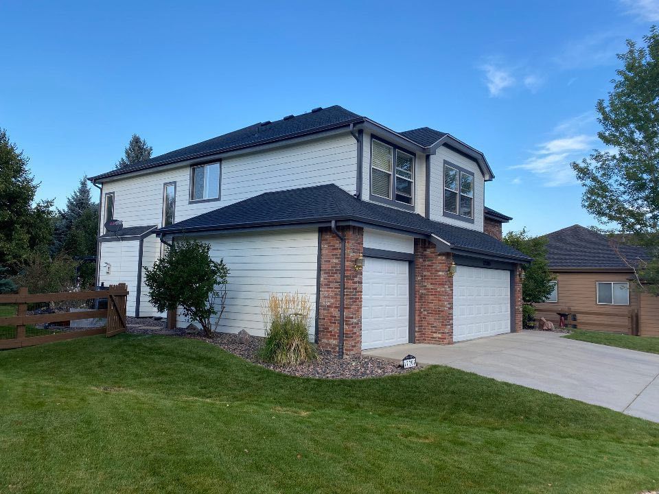 Two-story house with white siding, dark shingled roof, brick accents, and a double garage under a clear blue sky.