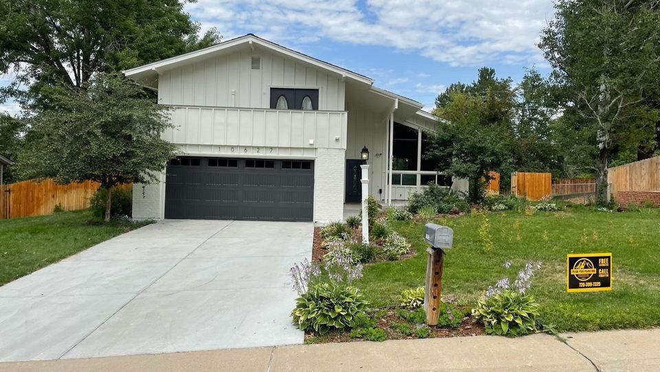 A white, two-story house with a black garage door, concrete driveway, and lawn with a real estate sign.