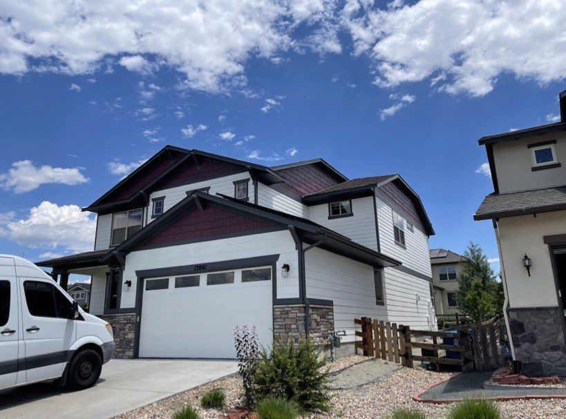 A two-story house with white siding, stone trim, and dark purple accents, viewed from the driveway on a sunny day.