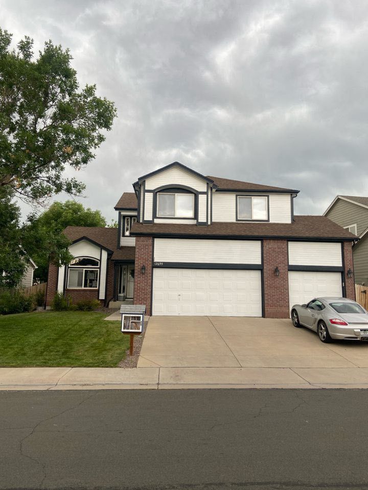 A two-story suburban home with white stucco, dark trim, and brick accents, featuring a three-car garage and a paved drive.
