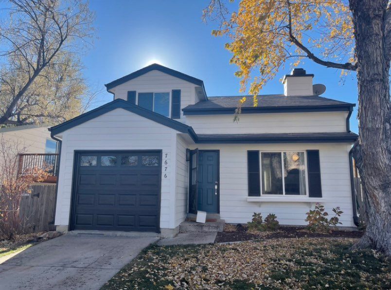 A two-story house with white horizontal siding, a black garage door, a front door, and a tree on a sunny day.