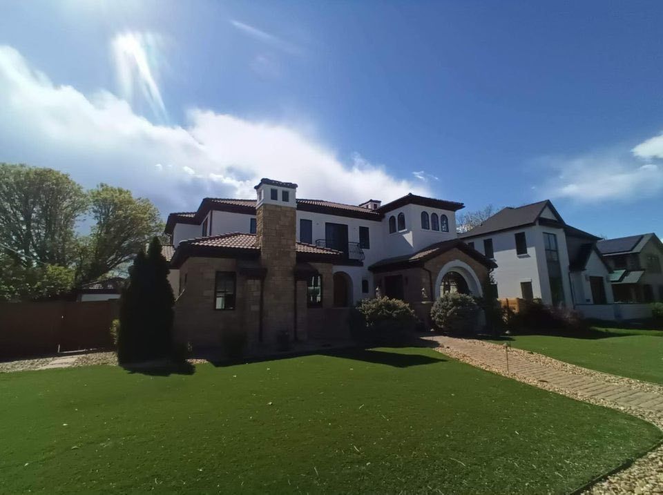 A two-story house with a tan stone facade, dark roof tiles, and a wide front lawn under a bright blue sky.
