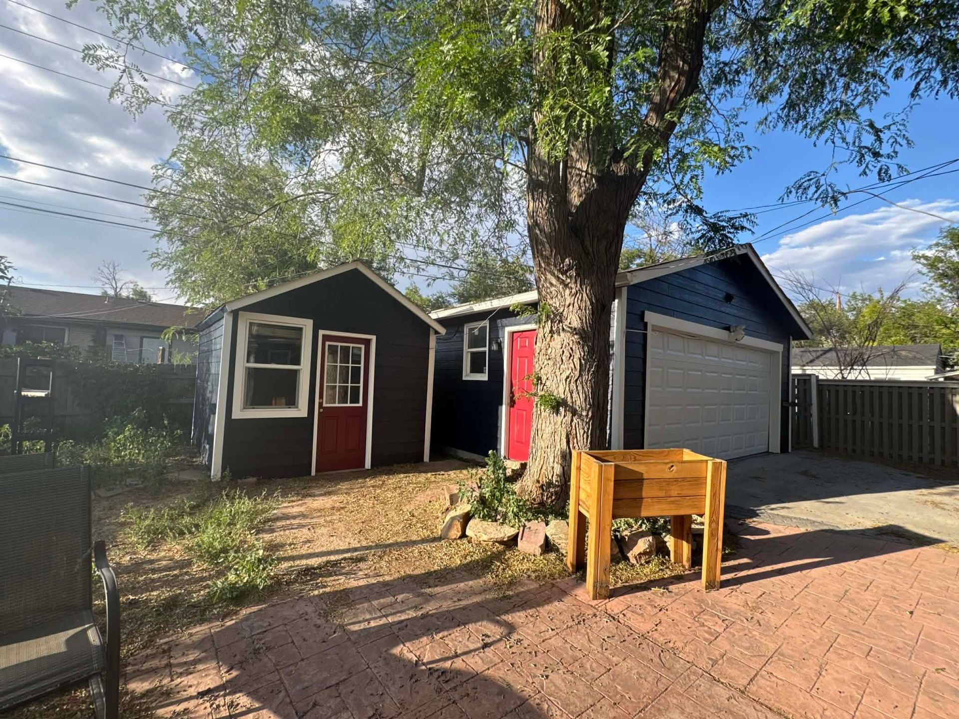 A dark blue shed and detached garage with red doors stand in a yard with a brick patio and a wooden planter box.