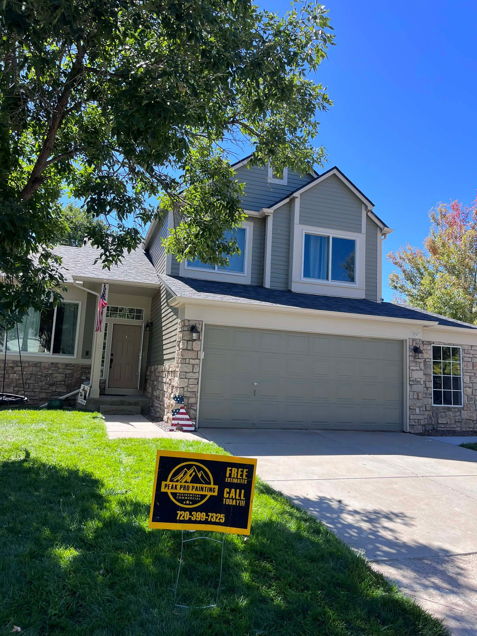 A two-story gray house with a garage and a brick exterior, featuring a lawn sign in the foreground under a blue sky.