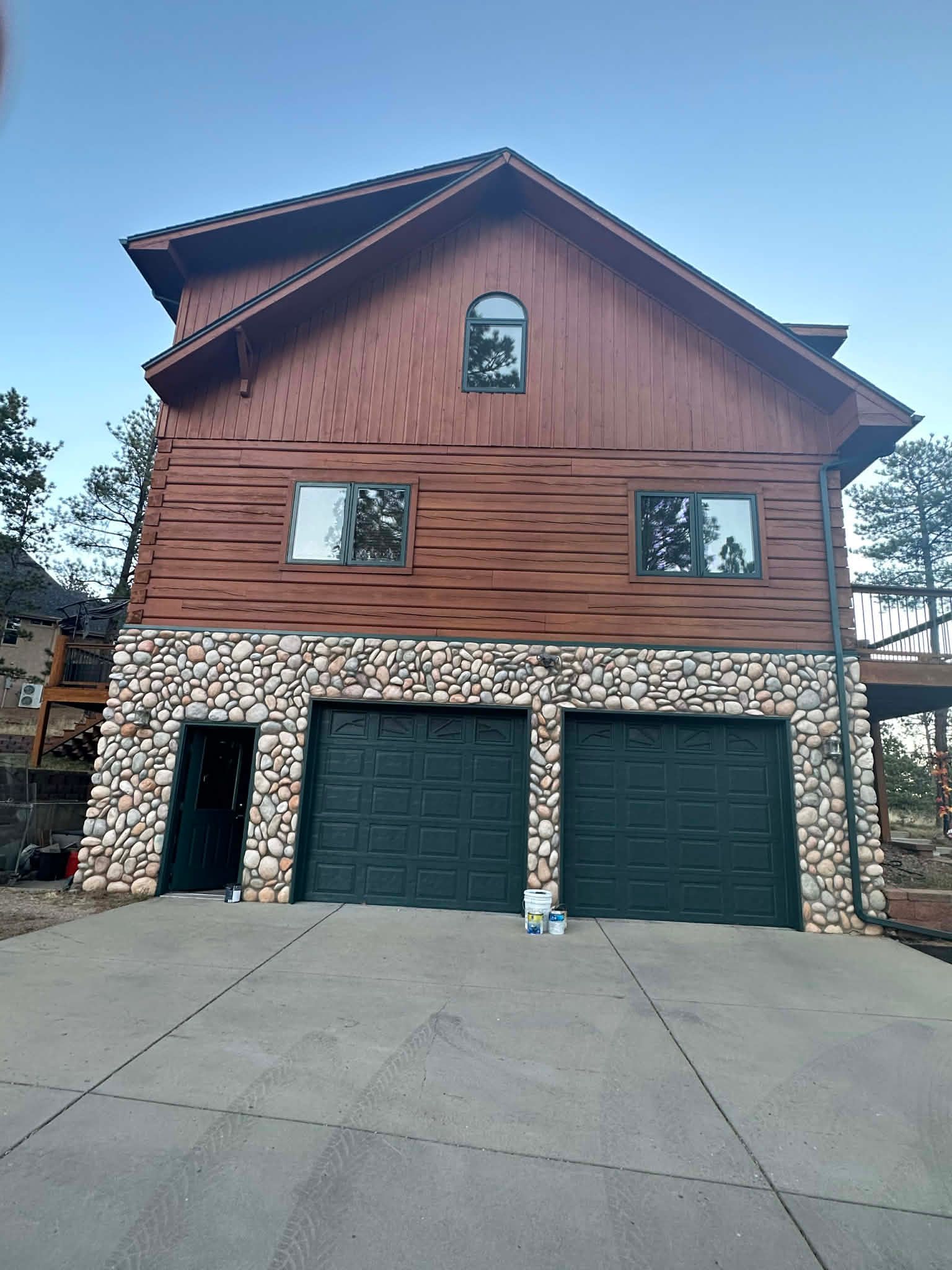 Two-story house with a rock-faced lower level, two green garage doors, and brown wood siding on the upper levels.