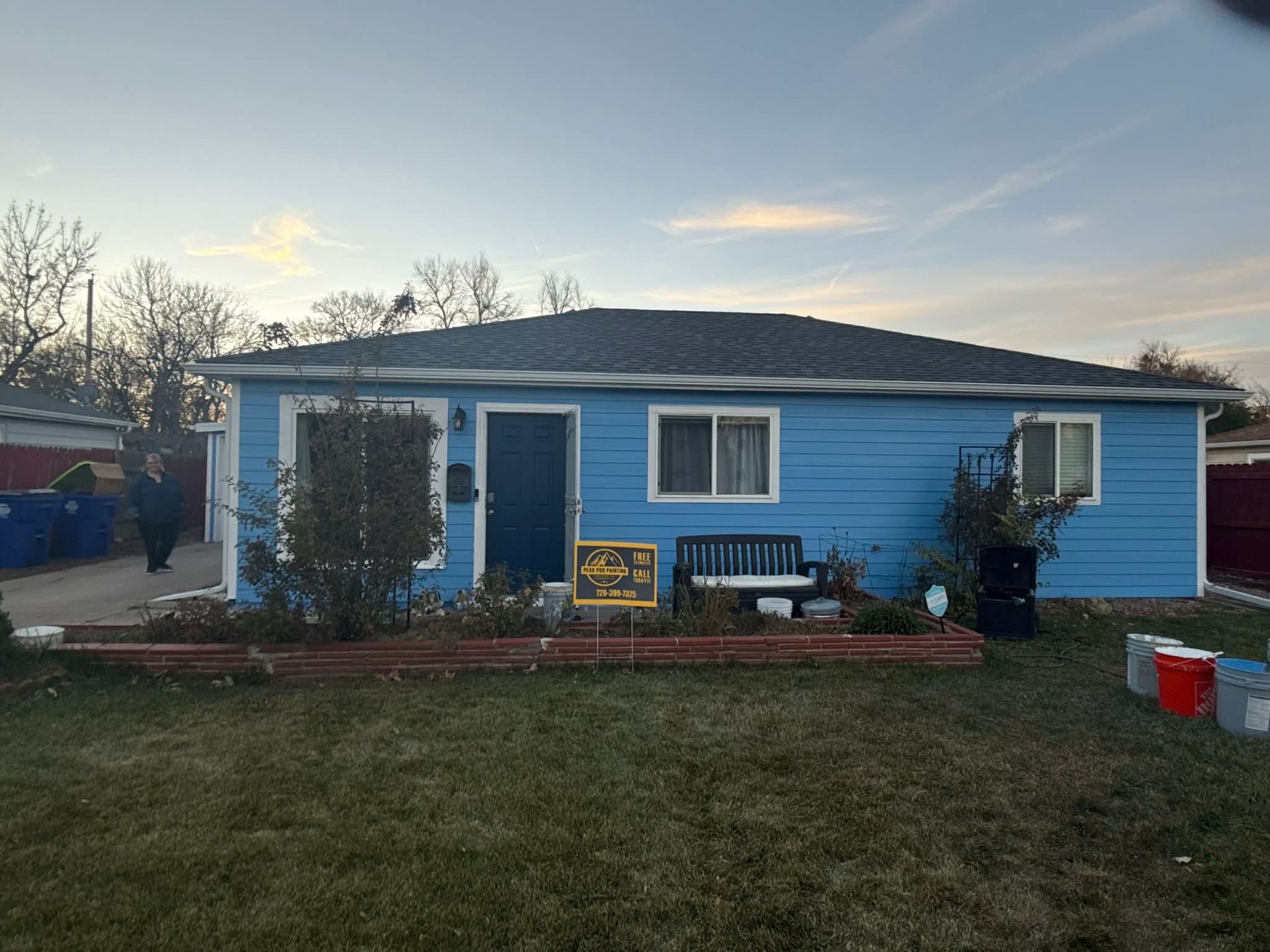 A single-story, bright blue house with a dark blue front door, white window frames, and a black bench in the front yard.