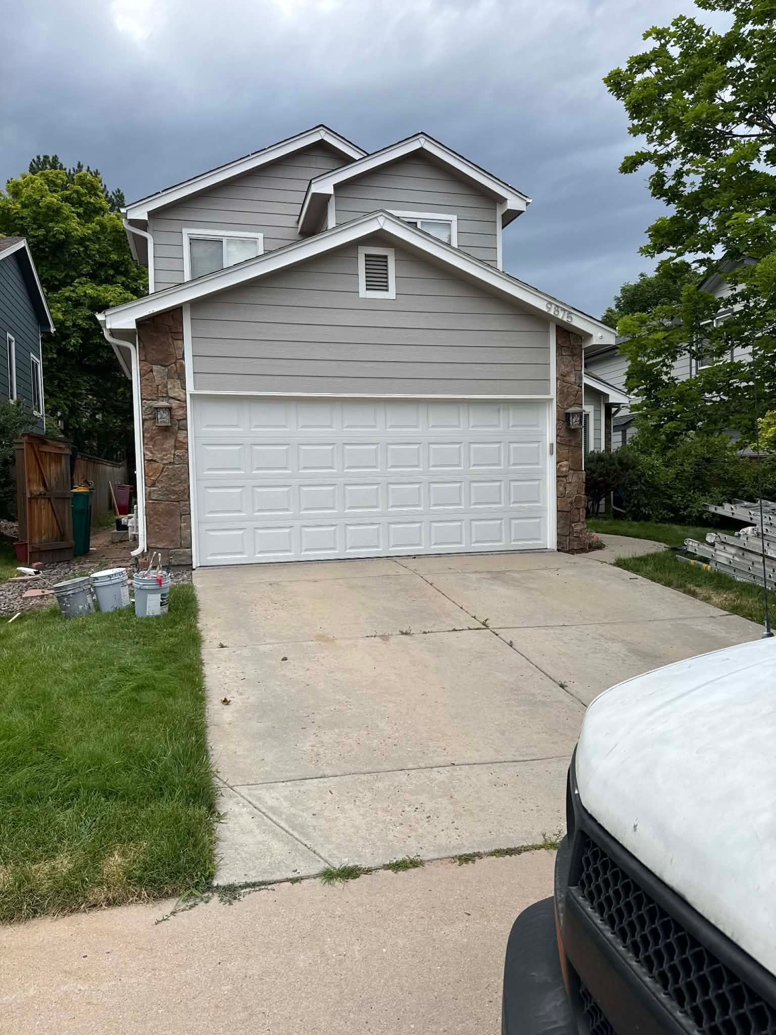 A two-story grey suburban house with a white garage door, a concrete driveway, and a partially visible vehicle in front.