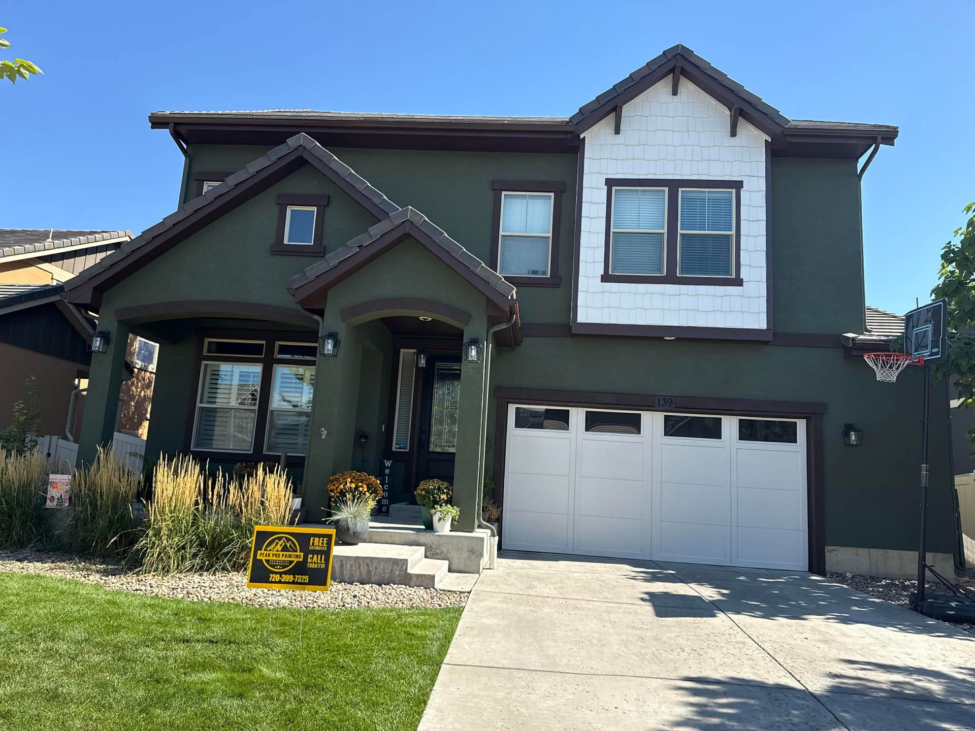 A two-story house with dark green stucco siding, a white garage door, and a brown shingle roof on a sunny day.